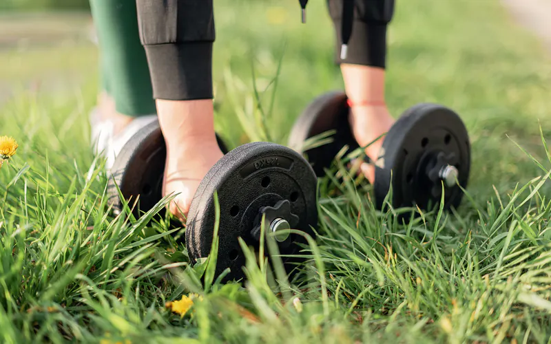 Hands gripping two plate dumbbells resting in long grass, shown from the shins down