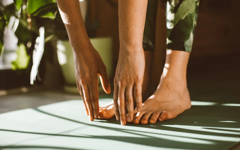 Hands reaching down to bare feet on a pale green mat in dappled sunlight