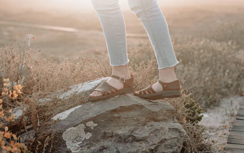 Feet in flat brown leather sandals stepping onto a rock among dry brush in warm evening light