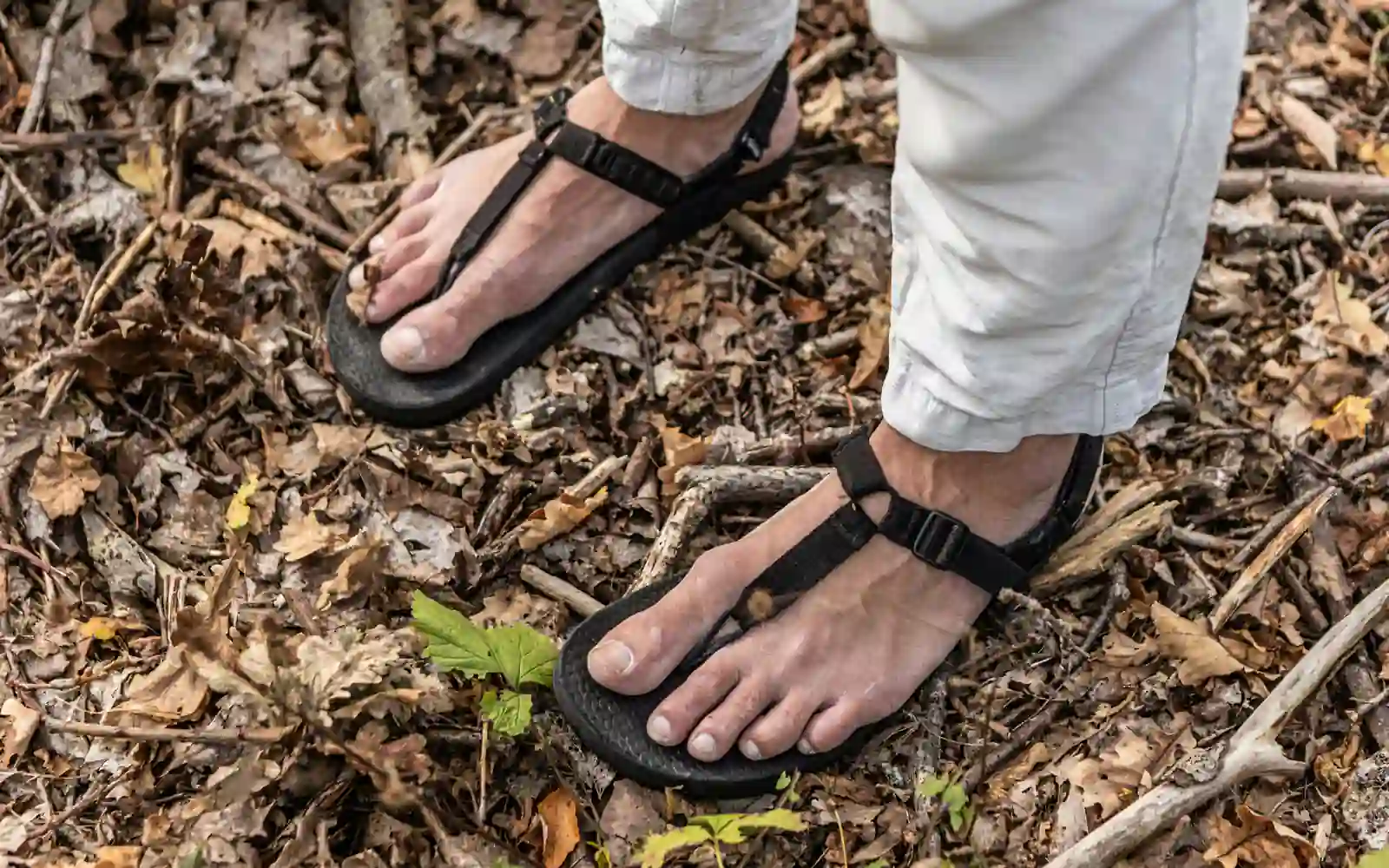 Feet in thin black strap sandals standing on a forest floor of dry leaves and twigs