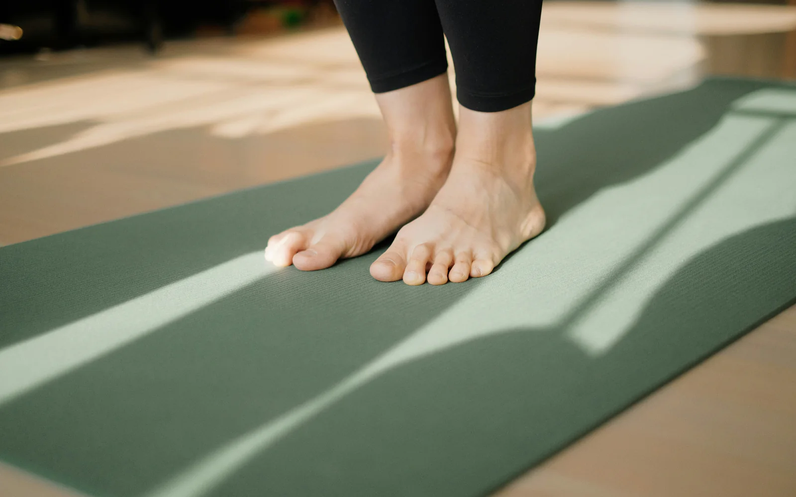 Bare feet standing on a sage-green mat on a wooden floor with bands of sunlight